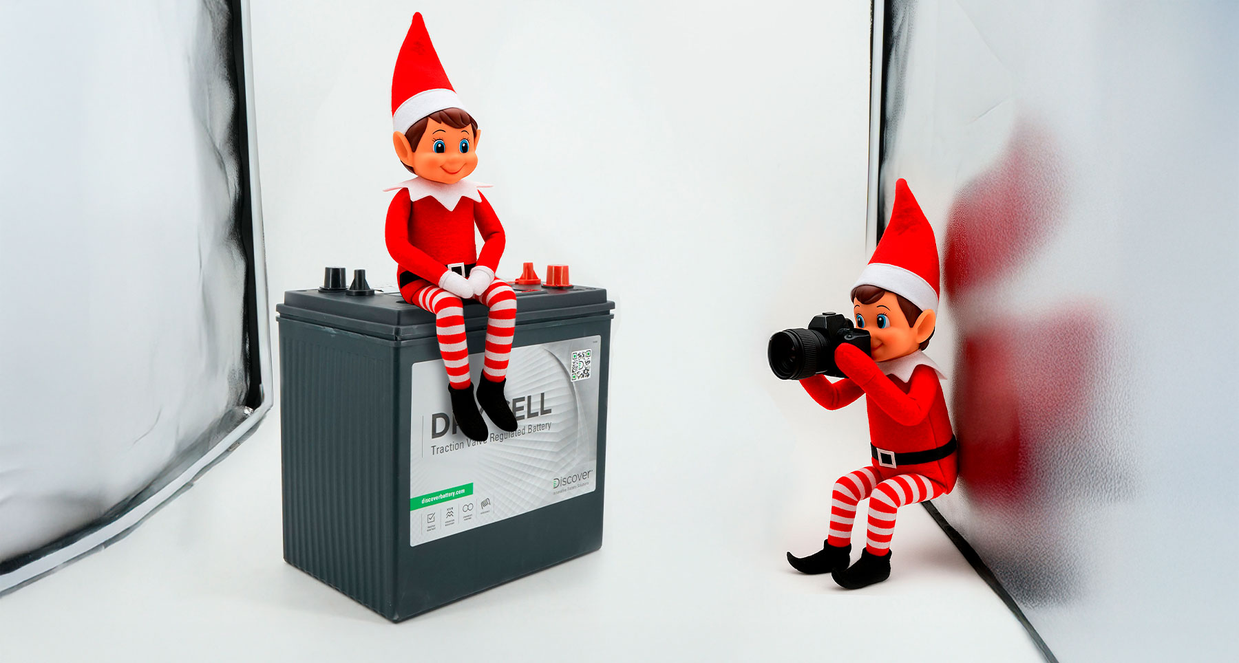 Dos elfos navideños haciendo una sesión de fotos sobre una batería AGM, uno posando y otro tomando fotos con una cámara.
